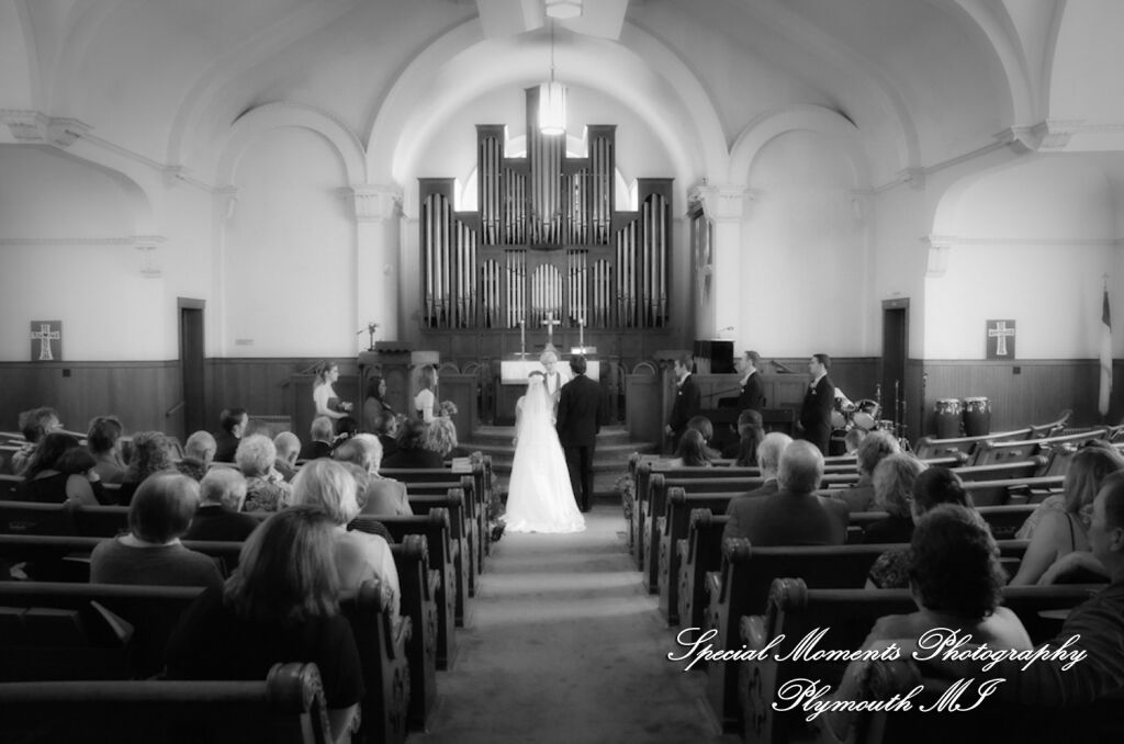 A wide-angle sanctuary photograph from the rear balcony captures a bride and groom exchanging vows at the altar before a majestic pipe organ inside the historic stone Chelsea First United Methodist Church in Chelsea, Michigan.