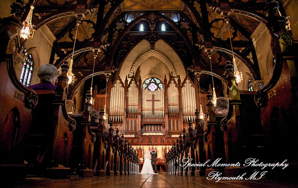 A wide-angle interior photograph from the back of the nave captures a bride and groom exchanging vows at the altar beneath the soaring Gothic Revival hammer-beam ceiling at Fort Street Presbyterian Church in downtown Detroit, Michigan.